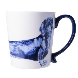 Op de afbeelding: Witte keramische mok met een blauw teckelontwerp. De hond is afgebeeld in een aquarel stijl. De mok heeft een donkerblauwe oor en een bijpassend blauw accent aan de basis van het oor.