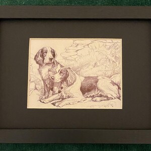 May include: A framed sepia-toned print featuring two dogs. One dog is seated, the other is lying down. The dogs have brown and white markings. The print is set in a black frame with a black mat.
