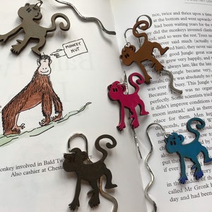 May include: A set of five monkey-shaped bookmarks, each with a different colour and design. The bookmarks are made of wood and have a silver metal wire attached to the top. The bookmarks are hanging from a book with the text "MONKEY NUT" visible.