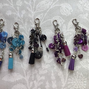 May include: Five keychains featuring a variety of coloured buttons, beads, and charms. Each keychain has a silver metal clasp and a tassel. The tassels are blue, black, and purple.