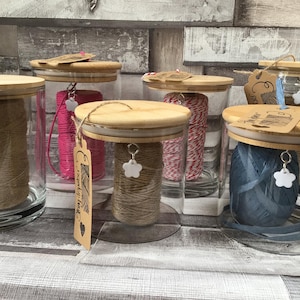 May include: Glass jars with wooden lids, filled with twine and string in shades of pink, blue, and natural tones. Each jar has a decorative tag and charm. The jars are displayed on a wooden surface.
