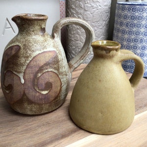 May include: Two ceramic jugs, one with a brown and white swirl design and the other a solid light brown. Both jugs have a handle and a spout.