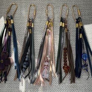 May include: Five keychains with gold-coloured clasps and decorative tassels. The tassels feature leather strips in various colours, including navy, brown, and pink, and are adorned with beads and a flower.