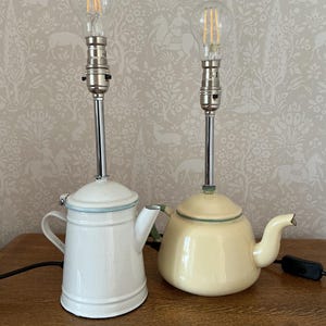 May include: Two table lamps fashioned from vintage enamel teapots. One lamp is a tall white cylindrical teapot, and the other is a shorter, rounded pale yellow teapot. Both feature chrome stems, silver-toned sockets, and clear filament bulbs.