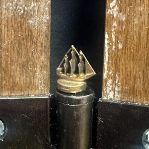 May include: A close-up of a gold-colored door hinge with a decorative ship design on the top. The hinge is attached to a wooden door frame.