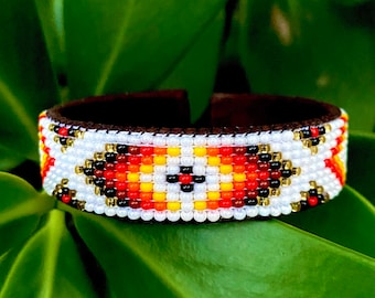 May include: A beaded bracelet with a geometric pattern in red, white, yellow, and black. The bracelet is on a green leafy background.