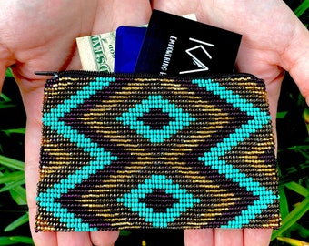 May include: A small beaded coin purse with a geometric pattern in turquoise, gold, and black. The purse is open and contains cash, a credit card, and a business card that reads "EMPOWERING A..."