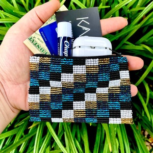 May include: A small, beaded pouch with a black, white, gold, and blue checkered pattern. The pouch is open and contains cash, a credit card, a lip balm, and a white case for wireless earbuds.