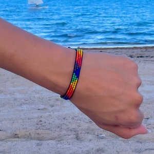 May include: A colorful beaded bracelet with red, orange, yellow, green, blue, and black beads. The bracelet is worn on a person's wrist against a background of a sandy beach and blue ocean.