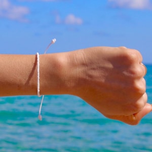 Könnte beinhalten: Ein weißes Perlenarmband mit einem weißen Schnurschluss. Das Armband wird am Handgelenk einer Person vor einem blauen Meereshintergrund getragen.
