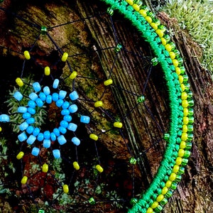 May include: A green, yellow, and blue beaded dreamcatcher with a black web. The dreamcatcher is hanging on a tree branch covered in moss.