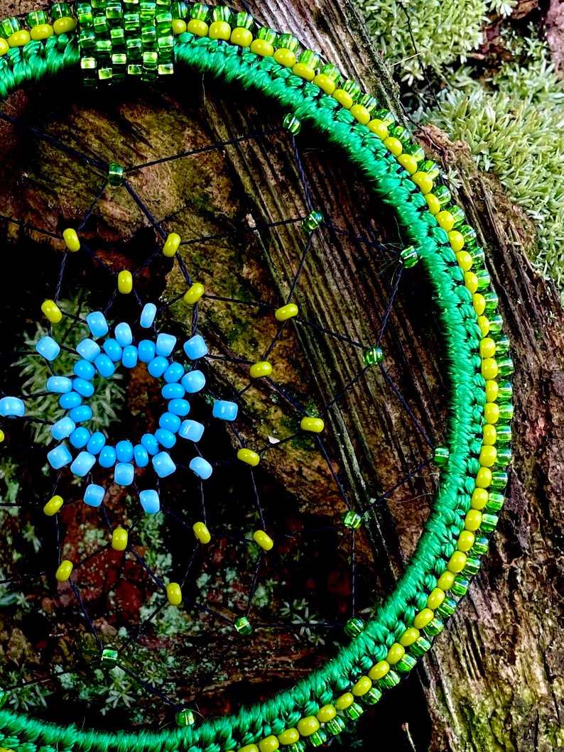 May include: A green, yellow, and blue beaded dreamcatcher with a black web. The dreamcatcher is hanging on a tree branch covered in moss.