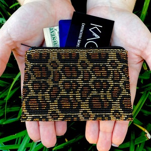 Puede incluir: Un monedero rectangular con cuentas y cierre de cremallera, con un diseño negro y dorado. El monedero contiene un billete de dólar, una tarjeta azul y una tarjeta negra con el texto "EMPOWERING HOME: KAC".