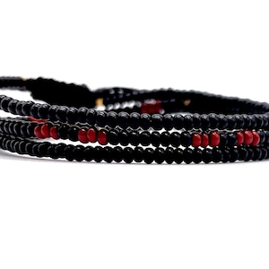 May include: Three black beaded bracelets with red accent beads. The bracelets are stacked on top of each other, with a braided cord closure. The beads are small and round, and the overall design is minimalist and modern.
