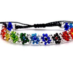 Op de afbeelding: Een regenboogkleurige kralen armband met een zwart koord en een verstelbare sluiting. De kralen zijn in een bloempatron gerangschikt en zijn rood, oranje, geel, groen, blauw, indigo en violet.