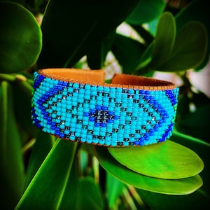 May include: A blue and green beaded bracelet with a brown leather base. The bracelet is made of small, colourful beads that are arranged in a geometric pattern.