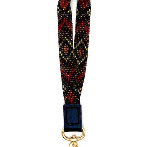 May include: A beaded wrist strap in a geometric pattern of red, black, and gold. The strap is attached to a navy blue leather piece and a gold-colored clasp. The strap is designed to be worn around the wrist.