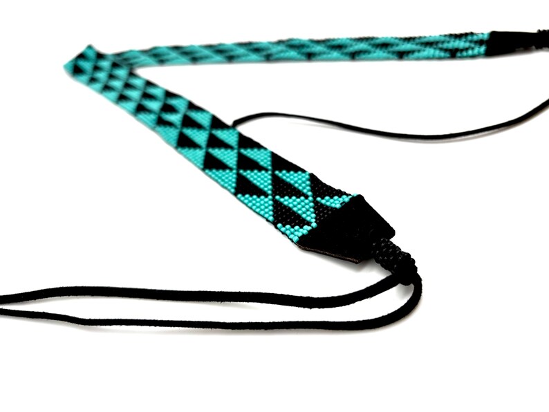 May include: A black and turquoise camera strap with a geometric pattern. The strap is made of woven beads and has a black leather adjustable strap.