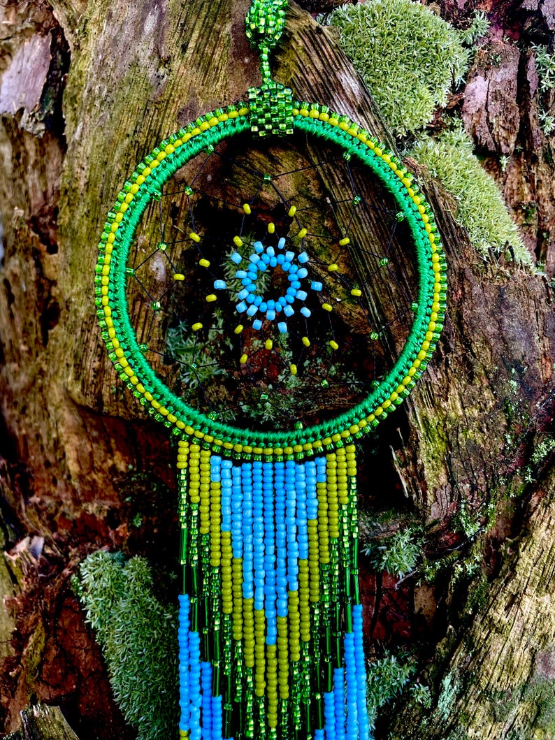 May include: A green, yellow, and blue beaded dreamcatcher with a circular frame and beaded fringe hanging from a tree trunk.