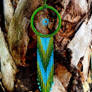 May include: A green and blue beaded dreamcatcher with a circular beaded frame and a long beaded fringe. The dreamcatcher is hanging from a tree trunk.