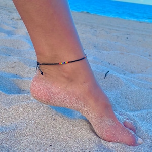 Op de afbeelding: Een zwarte kralen enkelband met gekleurde kralen op een zandstrand.