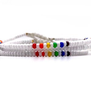 Puede incluir: Pulseras de cuentas blancas con un arcoíris de colores. Cada pulsera presenta una serie de pequeñas cuentas blancas con una sección de cuentas de colores en rojo, naranja, amarillo, verde, azul y morado. Las pulseras están superpuestas y tienen un cordón blanco.