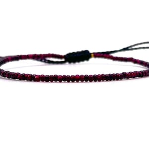 Könnte beinhalten: Ein zartes Armband aus kleinen, dunkelroten Perlen und einer schwarzen Schnur mit einem verschiebbaren Knotenverschluss.