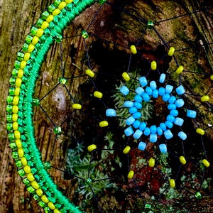 May include: A green and yellow beaded dreamcatcher with a blue and light blue beaded center. The dreamcatcher is hanging on a tree trunk.