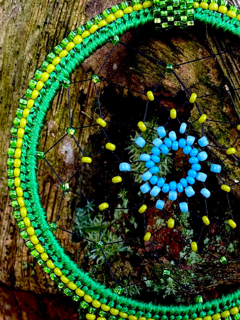 May include: A green and yellow beaded dreamcatcher with a blue and light blue beaded center. The dreamcatcher is hanging on a tree trunk.