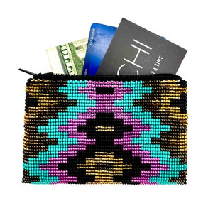 Puede incluir: Una pequeña bolsa con cremallera de cuentas con un patrón geométrico en turquesa, rosa, dorado y negro. La bolsa está abierta y tiene una tarjeta de crédito y dinero en efectivo en su interior.