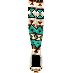 May include: A beaded wrist lanyard with a geometric pattern in turquoise, white, brown, and black. The lanyard has a black fabric connector and a gold-colored metal clasp. The design is symmetrical and features repeating shapes.