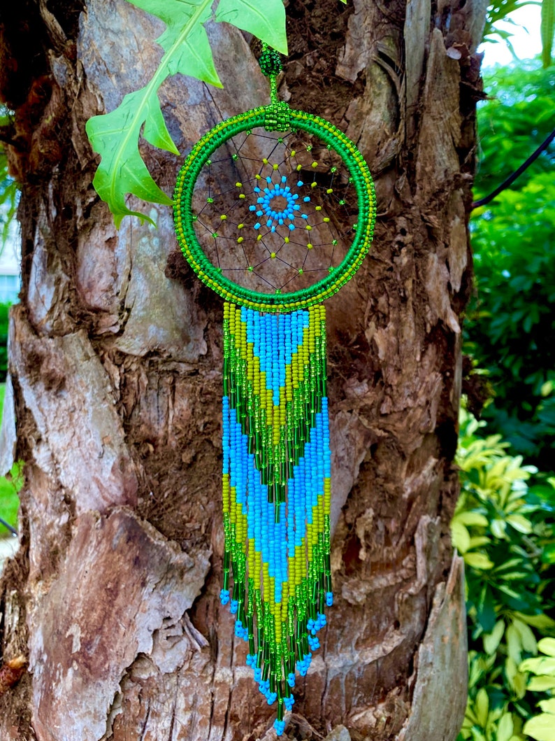 May include: A green and blue beaded dreamcatcher with a circular beaded frame and a long beaded fringe. The dreamcatcher is hanging from a tree branch.