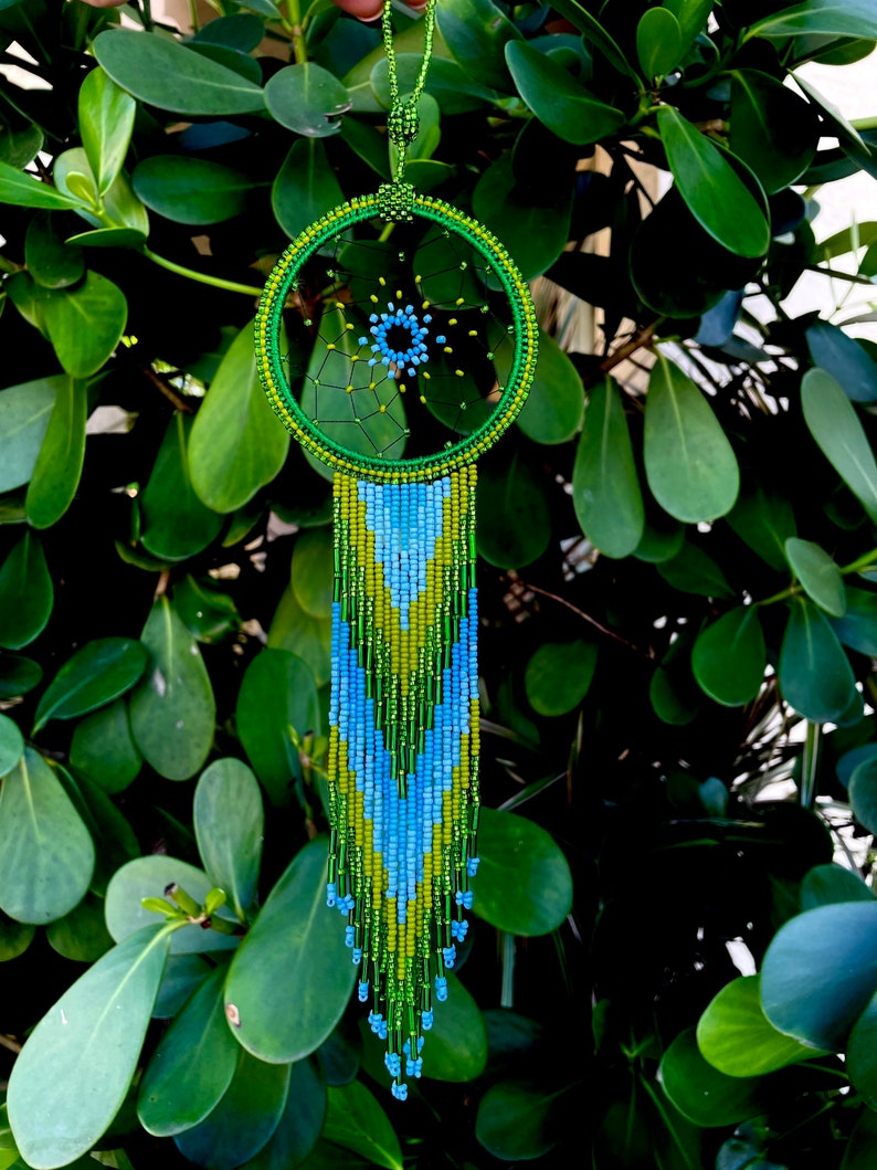 May include: Green, blue, and yellow beaded dreamcatcher with a fringe of beads. The dreamcatcher is hanging from a green cord.