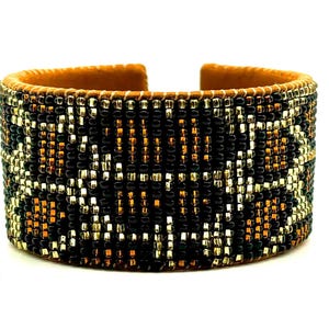 Könnte beinhalten: Breites Armband mit geometrischem Muster. Das Armband hat eine Innenseite aus beigem Wildleder und ist mit einem detaillierten Muster aus schwarzen, goldenen und bronzefarbenen Perlen verziert. Ein auffälliges Schmuckstück.