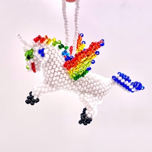 May include: A beaded unicorn ornament with a white body, rainbow wings, and a blue tail. The unicorn has a white horn and black hooves. The ornament is held by a white beaded string.
