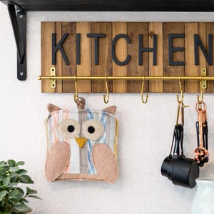 May include: A wooden sign with the word "KITCHEN" in black letters hangs above a brass rod with hooks. An owl-shaped pot holder with tan, blue, and peach colors hangs below the sign. Kitchen utensils hang from the hooks.