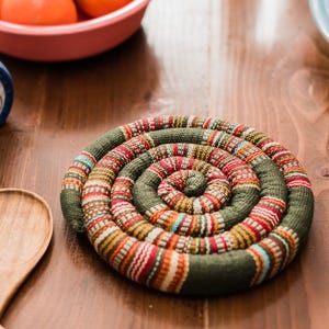 May include: A green and orange woven pot holder with a spiral design. The pot holder is on a wooden table with a wooden spoon and a blue and white ceramic vase.