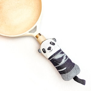 Puede incluir: Una sartén de cerámica blanca con un agarrador de olla con temática de panda en blanco y negro. El agarrador de olla tiene un patrón de rayas blancas y negras y una cara de panda con ojos negros y una nariz negra.