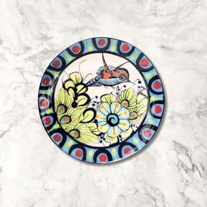 May include: A blue and white ceramic plate with a red and yellow border. The plate features a hand-painted design of a hummingbird flying over a cluster of flowers.