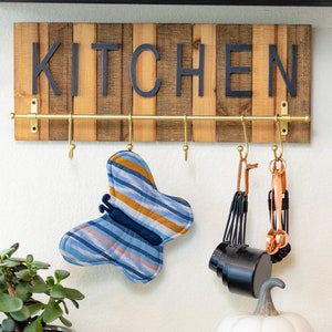 May include: A wooden sign with the word "KITCHEN" in black letters hangs above a gold metal rack with hooks. A blue and yellow striped pot holder hangs from one hook, and two sets of measuring spoons hang from other hooks.