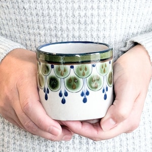 May include: A ceramic mug with a white base, adorned with a repeating pattern of green and brown teardrop shapes, outlined in blue. The rim is trimmed with a dark green band and a blue inner rim. The mug is held in both hands.