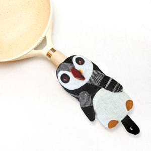 Puede incluir: Un protector de mango de olla con forma de pingüino en blanco y negro, con pico rojo y patas marrones. El protector está sujeto a una sartén color crema con un mango de madera. El pingüino tiene ojos grandes y redondos y es de tela.