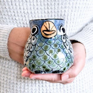 Puede incluir: Una taza de cerámica azul y verde con forma de pájaro con una cara sonriente. La taza tiene una superficie texturizada que se asemeja a escamas.