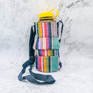 May include: A colorful striped fabric water bottle carrier with a black adjustable strap. The carrier has a clear plastic water bottle inside with a yellow lid.