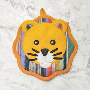May include: A whimsical, handmade lion-shaped pot holder. The lion's face is a bright yellow with black whiskers and eyes, and a white muzzle. The pot holder has a colorful striped fabric backing and an orange border. A loop is attached for hanging.