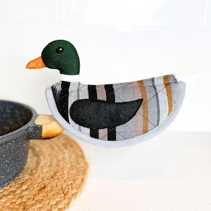 Puede incluir: Un agarrador de olla a rayas grises y marrones con forma de pato con un cuerpo de fieltro negro. El pato tiene una cabeza verde y un pico naranja.