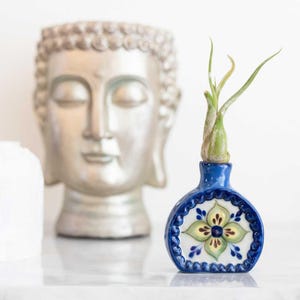 May include: A small, blue ceramic vase with a floral design, holding a green air plant. The vase has a rounded shape and a detailed, hand-painted pattern in white, green, and blue. A silver Buddha head is in the background.