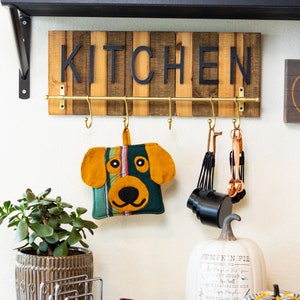 May include: A wooden sign with the word "Kitchen" in black letters hangs above a gold metal rack with hooks. A green and yellow dog-shaped pot holder hangs from one of the hooks.