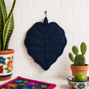 Puede incluir: Un agarrador de tela azul marino oscuro con forma de hoja grande y costuras acolchadas decorativas que imitan las venas de una hoja. El agarrador cuenta con una pequeña presilla de tela en la parte superior para colgarlo en una pared de azulejos blancos.