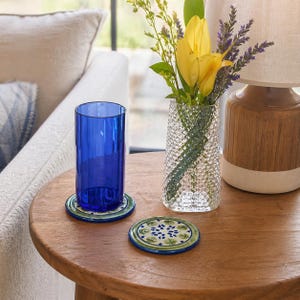May include: A blue glass tumbler sits on a decorative coaster with a green and white floral design. Another coaster is next to it. A clear textured vase holds yellow tulips and purple flowers. A wooden lamp is in the background.